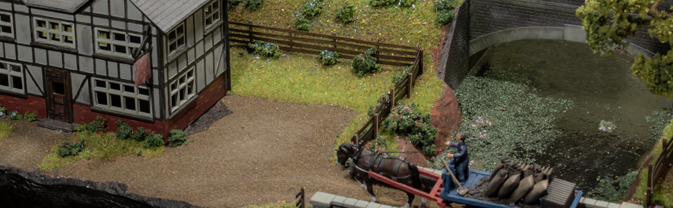 Miniature model scene featuring a traditional half-timbered building with surrounding landscape and water feature.