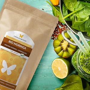 Sachet en papier brun contenant de la poudre de Moringa bio, entouré de fruits frais, de légumes et d'un smoothie vert sur fond turquoise