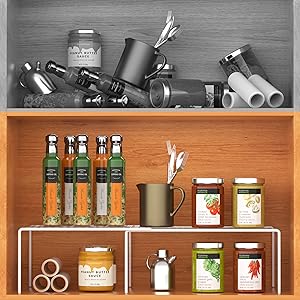 Two-tiered wooden shelf organizer displaying various kitchen items. Lower shelf holds glass jars and bottles, upper shelf contains metal utensils and cookware.