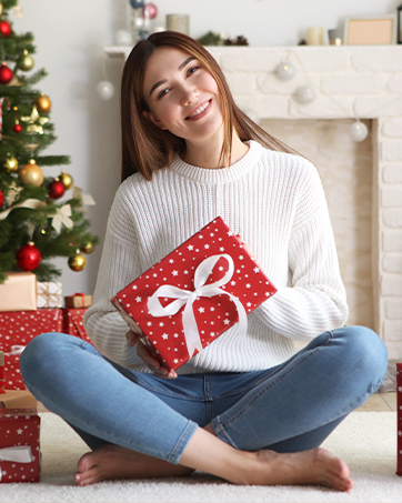 Woman in white sweater sitting on floor, holding red gift box with white bow. Christmas tree visible in background.