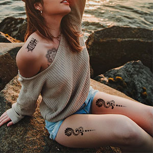 Persoon die op een rotsachtige kust zit en een off-shoulder trui en korte broek draagt. Donkere bloementattoos zichtbaar op dij en bovenarm.