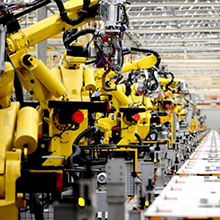 Multiple large yellow industrial robotic arms in a manufacturing facility. The robots are arranged in a row, suggesting an automated assembly line setup.