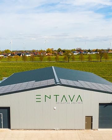 Industriegebäude mit grauem Metalldach und „ENTAVA“-Schild vor einer ländlichen Landschaft mit grünen Feldern.