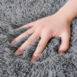 A hand resting on a plush, gray shag carpet or rug. The fingers are spread, emphasizing the soft, dense texture of the carpet fibers.