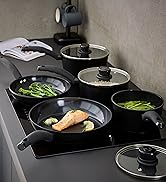 Multiple black non-stick pans on stovetop cooking various foods including green vegetables and protein, with glass lids visible.