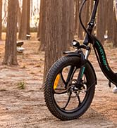 Primer plano de la rueda delantera y parte del cuadro de una bicicleta negra en un entorno forestal con árboles visibles al fondo