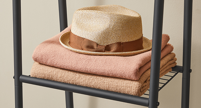 Bézs színű szalmakalap barna szalaggal, összehajtogatott barackszínű törölközőkön, fémpolcon.