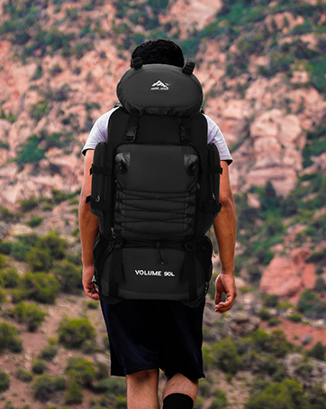 Grand sac à dos noir porté par le randonneur face à la caméra. Scène extérieure accidentée avec des formations rocheuses rouges en arrière-plan.