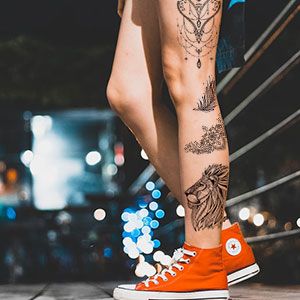 Oranje hoge sneakers gedragen op voeten met zichtbare tatoeages op de benen. Nachtelijke stadsomgeving met wazige lichten op de achtergrond