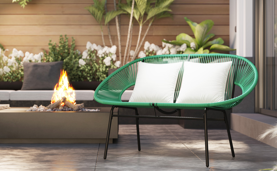 Cadre de terrasse extérieur avec une causeuse tissée verte, des coussins blancs et un petit foyer. La causeuse a un design incurvé et des pieds en métal noir