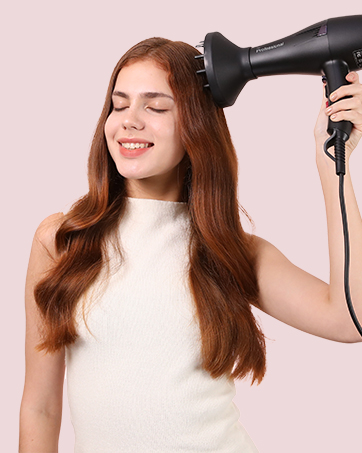 Black hair dryer in use, drying long auburn hair. Compact design with visible nozzle attachment. Person wearing white sleeveless top against pink background.
