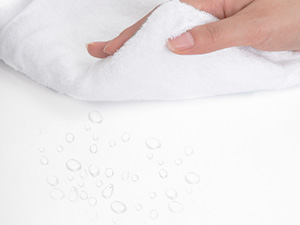 Nahaufnahme einer weißen Stoffoberfläche mit Wassertröpfchen. Eine Hand wird gezeigt, wie sie das Material berührt und dessen wasserabweisende Eigenschaften demonstriert