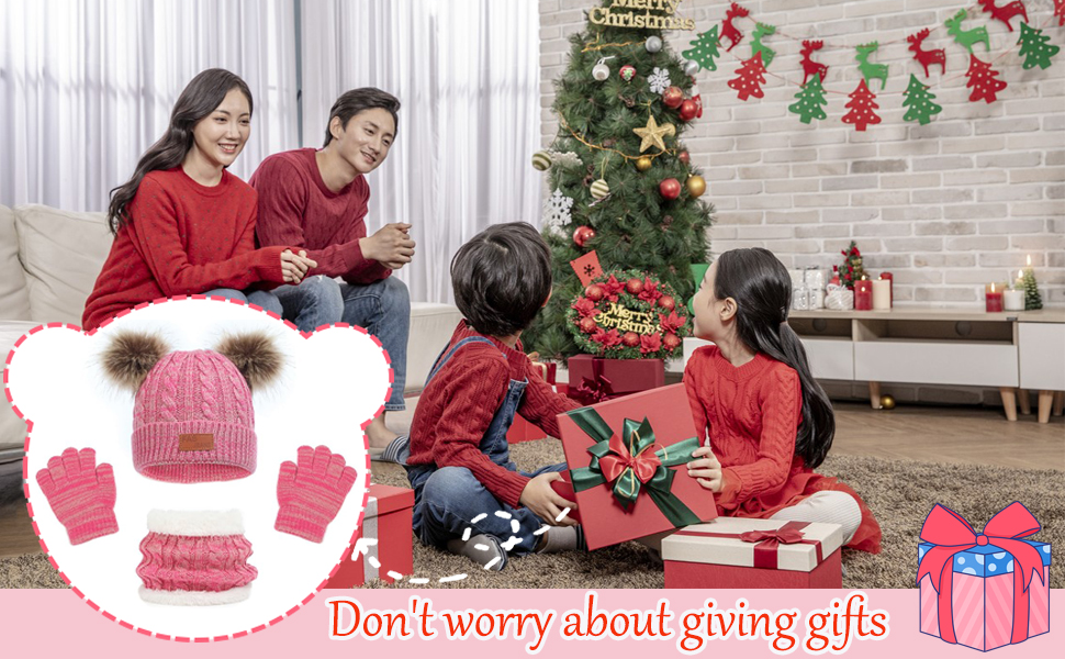 Pink winter accessory set with hat, scarf, and gloves. Hat features fur pom-poms. Set displayed against Christmas family scene background.