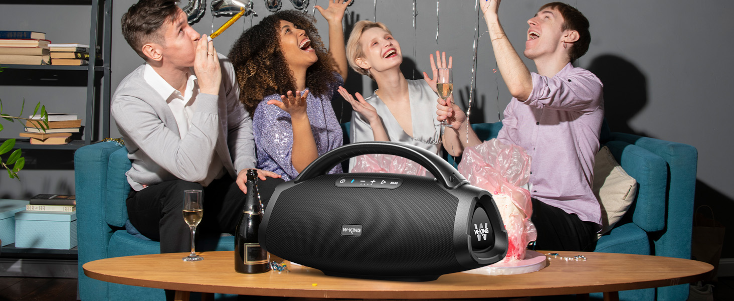 Large portable speaker on coffee table with group enjoying music. Black cylindrical device with handle, surrounded by people raising glasses in celebration.