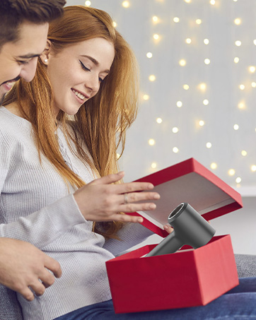 Deux personnes ouvrent un coffret cadeau rouge, révélant un article gris à l'intérieur. Des guirlandes lumineuses festives sont visibles en arrière-plan.