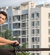 Una persona apoyada en el manillar de una bicicleta en un balcón, con edificios de apartamentos visibles al fondo