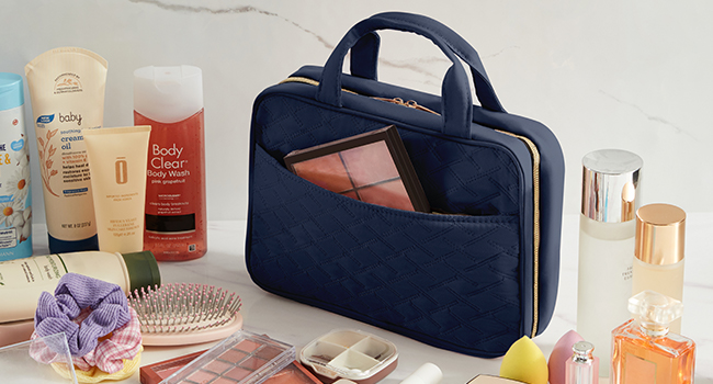 Navy blue toiletry bag surrounded by various beauty products including lotions, perfumes, and a hairbrush. Bag has a zippered main compartment and carrying handles.