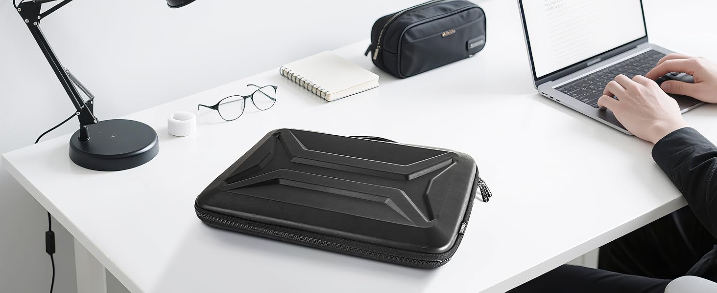 Modern desk setup with laptop, desk lamp, and black protective case. White desk surface with minimal workspace arrangement.