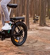 Bicicleta eléctrica con neumáticos gruesos en un sendero forestal. Se ven las piernas del ciclista, mostrando la bicicleta en uso. Marco negro con detalles en naranja.