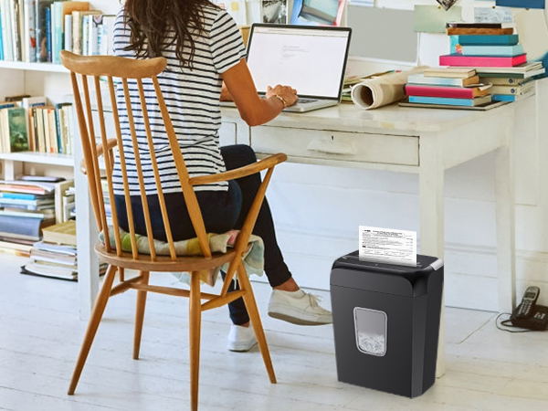 Paper shredder placed under a white desk in a home office setting. Wooden chair and bookshelves visible in the background.