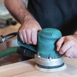 A hose from a shop vacuum is attached to a handheld orbital sander working on smoothing wood