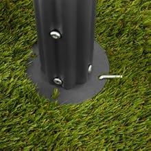 Gros plan d'une base de poteau en métal noir sur du gazon artificiel. Le mât est fixé à une plaque métallique circulaire à l'aide de vis visibles et d'une goupille de verrouillage.