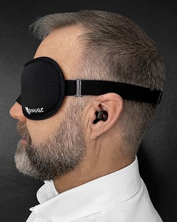Black sleep mask with adjustable strap worn by a person. Includes a small earbud or hearing device visible in the ear.