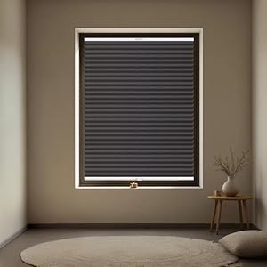Blackout cellular window shade installed in a minimalist room, featuring horizontal pleated design and bottom pull handle. Round beige rug and wooden side table visible below.