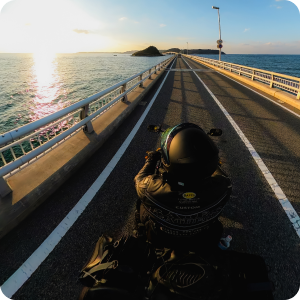 Vista en primera persona desde una motocicleta en un puente durante el atardecer o el amanecer. El océano es visible desde ambos lados, con barandas metálicas a lo largo del puente y marcas viales claramente visibles.