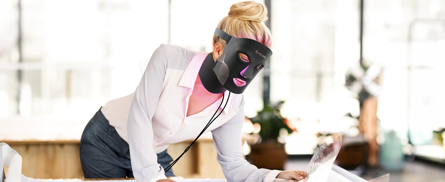 Black facial massage device with pink accents being used on cheek. Electric cord visible. User wearing light-colored top in bright indoor setting.