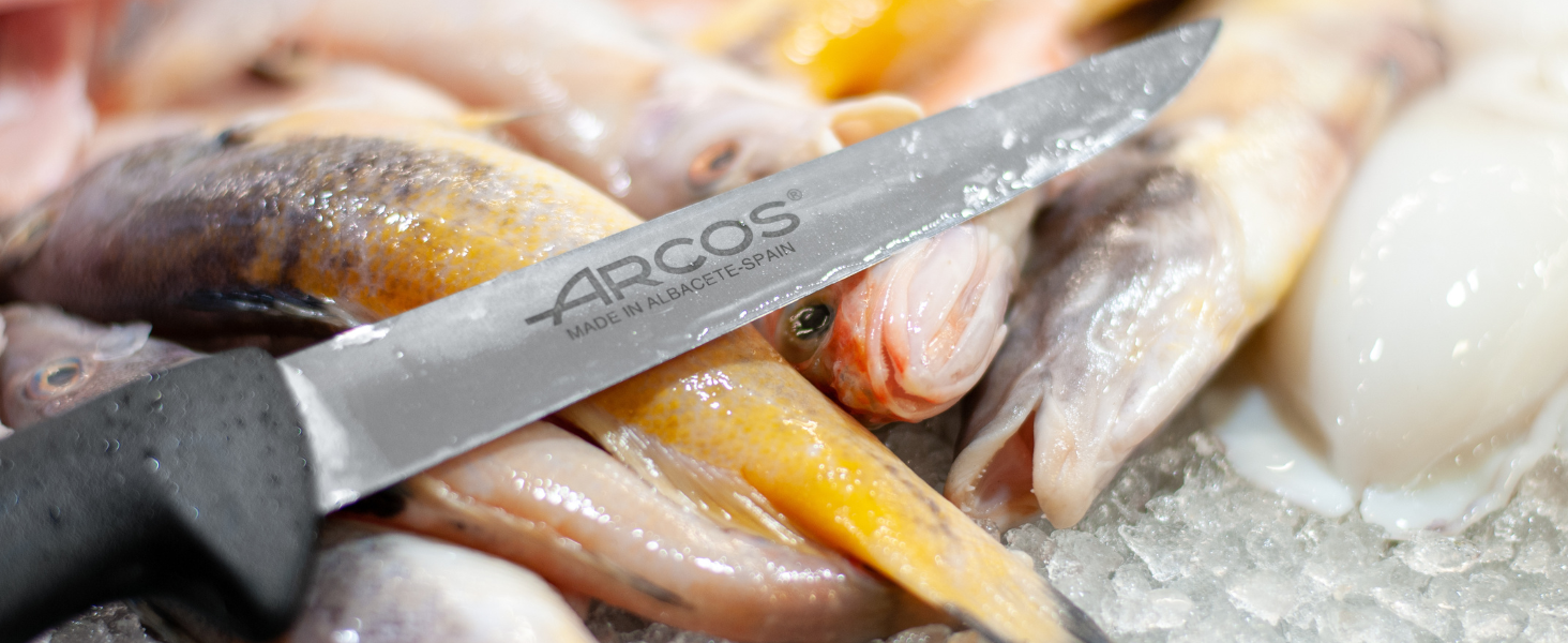 Primer plano de pescado crudo cortado con un cuchillo. La hoja con la etiqueta «Arcos» es visible, lo que sugiere un contexto profesional de cocina o preparación de alimentos