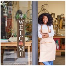 A woman in apron smiles by a glass door with a &#34;WELCOME&#34; sign and maple leaf.