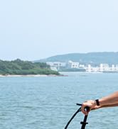 Mano que sostiene una caña de pescar con carrete, extendida sobre el agua. Paisaje costero con edificios visibles en segundo plano.