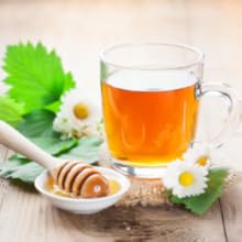 Honey and tea service presentation with chamomile flowers, honey dipper, and amber-colored tea in glass cup on light wooden surface.