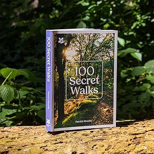 Text reads '100 Secret Walks'. Book cover photographed outdoors showing nature path through trees, placed on wooden surface with greenery in background.