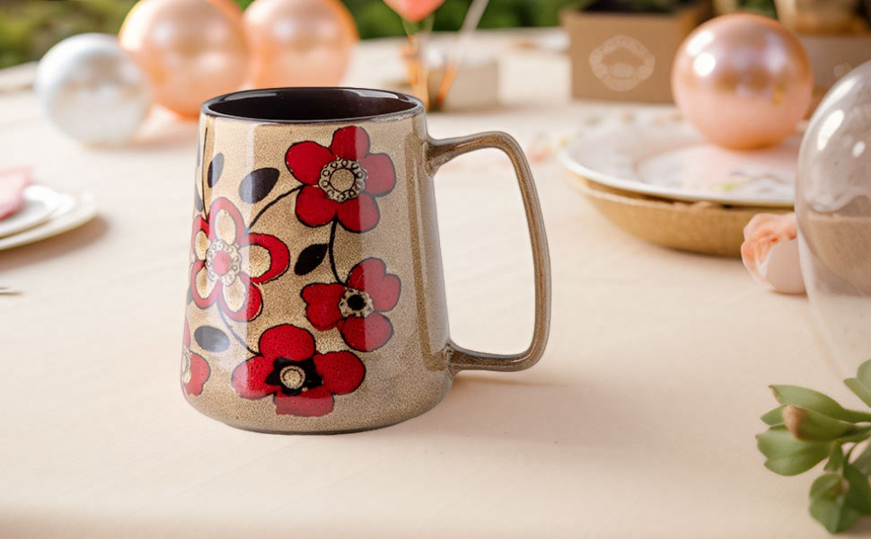 Ceramic mug with tapered design featuring red and pink floral pattern on beige background, displayed on table with decorative settings.