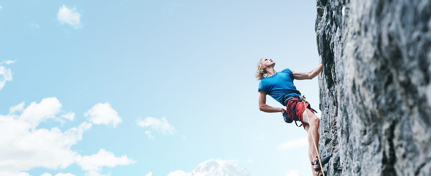 Escalador con camisa azul y arnés rojo que escala un acantilado empinado contra un cielo azul con nubes