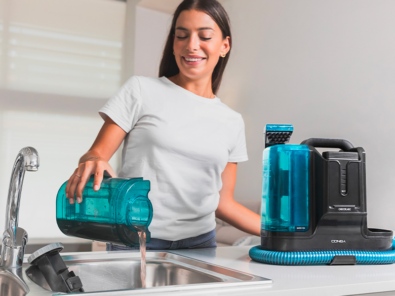 Cecotec Conga Carpet&Spot Clean Infinity Cordless Upholstery and Carpet Vacuum Cleaner. 150W, 2 Interchangeable Batteries, 60Mins Autonomy, Water Sprayer and Window Cleaner Accessory 17 Mujer que usa un limpiador a vapor portátil de color verde azulado en el fregadero de la cocina, con un dispositivo de limpieza adicional en la