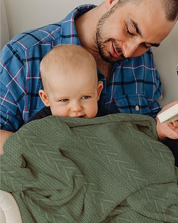 A person in a blue plaid shirt holding a baby wrapped in a green blanket. They are looking down, seemingly engaged with something out of view.