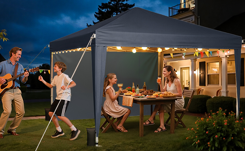 A large outdoor canopy tent with LED lighting illuminating a dining area at dusk, set up on a lawn with garden furniture underneath.