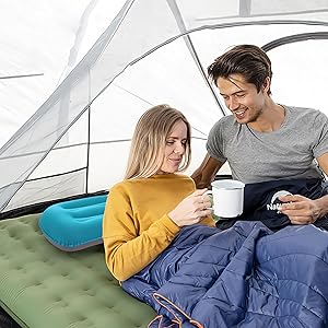 Scène de camping avec deux personnes dans des sacs de couchage sur un matelas gonflable à l'intérieur d'une tente. Ils boivent dans des tasses et du matériel de camping supplémentaire est visible.