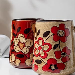 Two decorative ceramic cups with floral designs featuring red and beige flowers with black accents, one in red base color and one in beige.