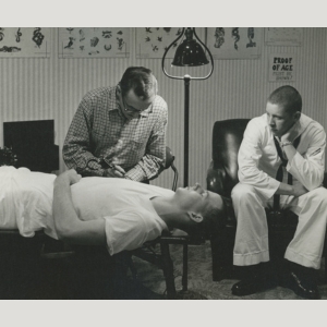 Black and white photo of a man giving another man a tattoo on his arm, while a third man watches.