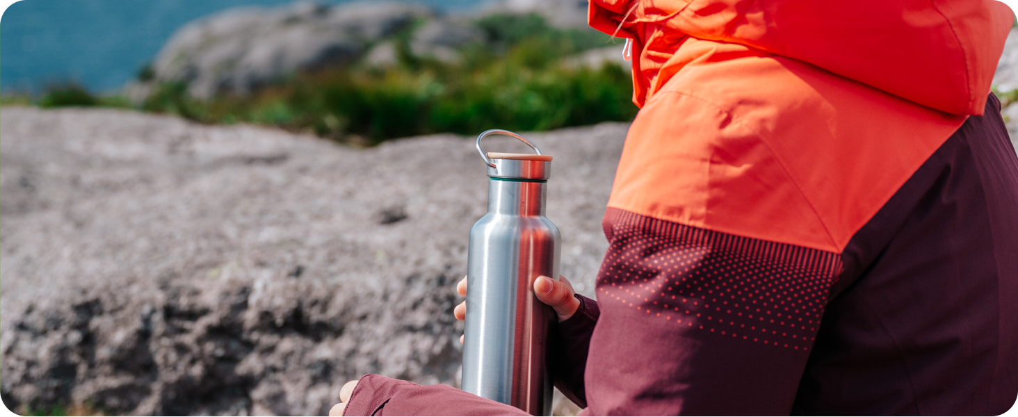 Stainless steel water bottle being held against an outdoor rocky background, with someone wearing orange and burgundy clothing.