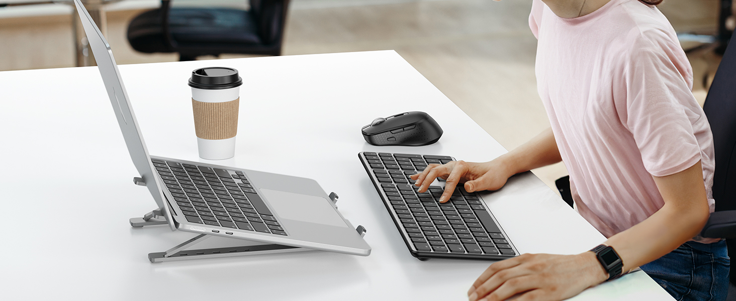 Two laptops on white desk with elevated laptop stand and coffee cup visible. Workspace setup showing ergonomic positioning of devices.