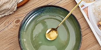 Green ceramic bowl containing liquid on wooden surface. Gold spoon in bowl, surrounded by plates with food and utensils.