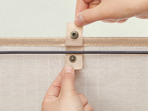 Close-up of hands fastening a beige snap button closure on a fabric surface, likely securing a zipper or joining two fabric panels.