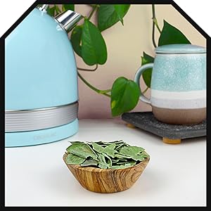 Bol en bois rempli de feuilles vertes séchées, bouilloire bleue et tasse en céramique sur surface blanche. Plante en pot avec des feuilles en forme de cœur