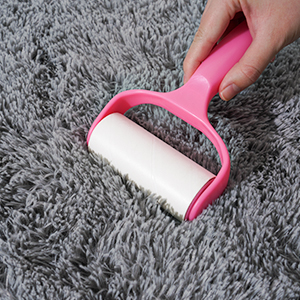 Pink handheld lint roller being used on gray fabric or carpet. Roller has white adhesive surface for collecting debris.