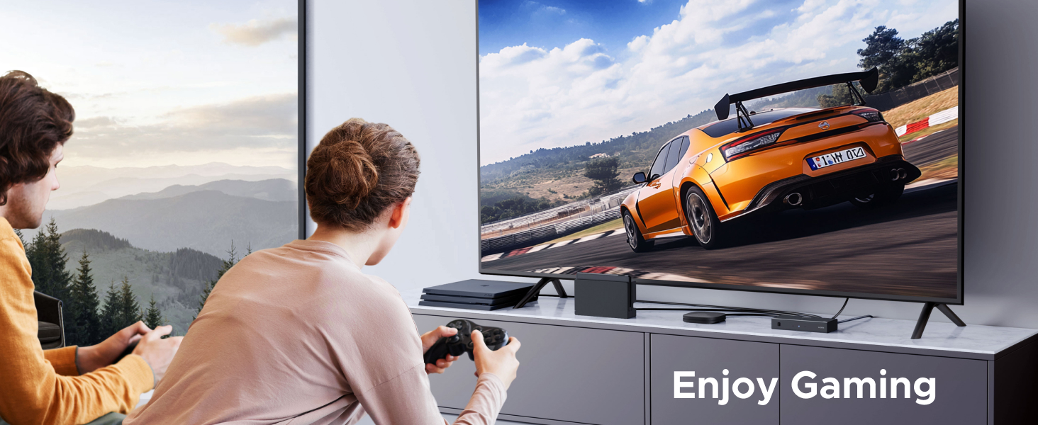 Large flatscreen TV displaying a racing video game with an orange sports car on a track. Two people sit on a couch holding game controllers, playing the game.